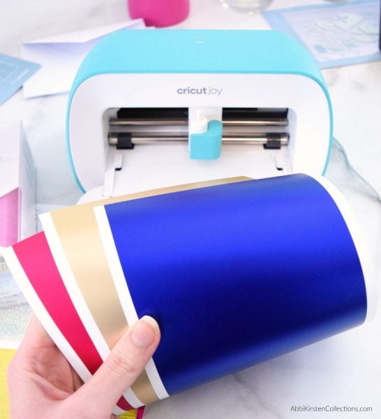 Abbi holds pink, gold and blue Cricut Smart Vinyl sheets in front of a blue Cricut Joy.