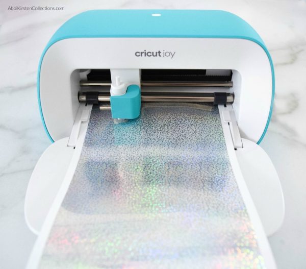 A long holographic smart vinyl sheet is being loaded into a Cricut Joy. 
