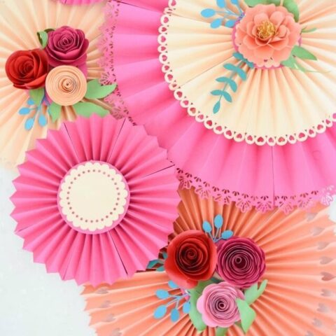 Pink and peach paper rosette fans hang overlapped on a white wall, with paper roses and greenery adorning the center of the decorations.