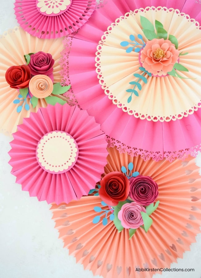 Pink and peach paper rosette fans hang overlapped on a white wall, with paper roses and greenery adorning the center of the decorations. 