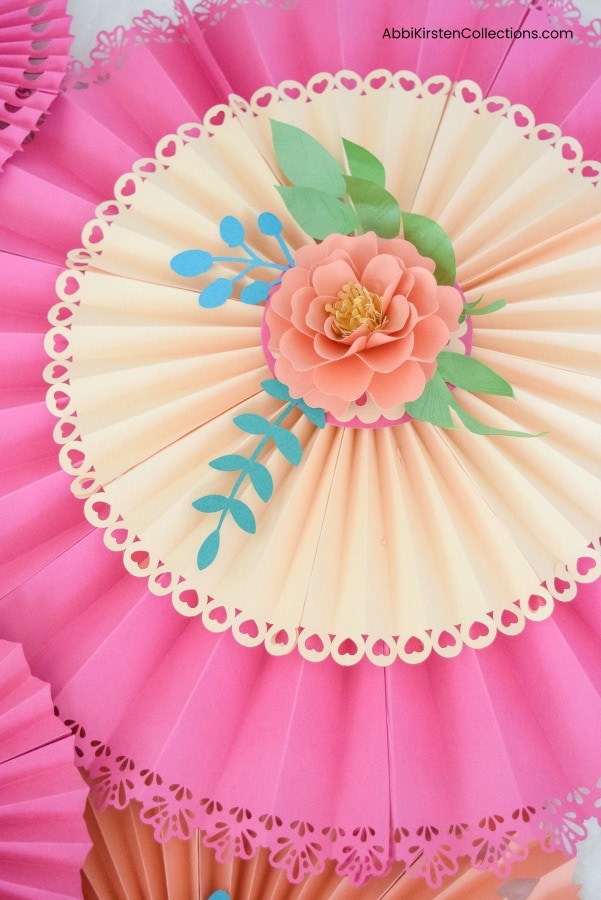 A closeup of an orange paper flower with green and blue leaves atop a vanilla and pink paper rosette fan. You can make these with Abbi Kirsten's template and video instructions.