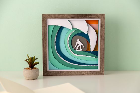 A framed craft made from layered cut paper sits on a table top next to a small succulent plant. The layered paper makes an ocean wave and a surfer. 