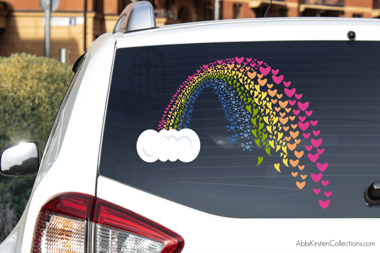 A rainbow SVG window cling made up of colorful hearts pouring out of a cloud on the back window of a white car.