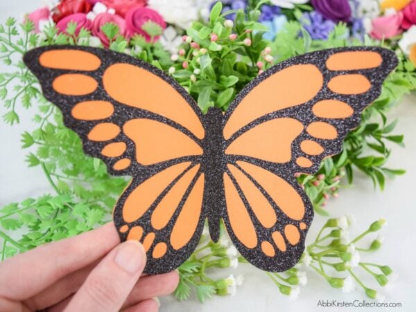 A woman's hand holds a black and orange finished butterfly craft using vinyl on cardstock. These 25 vinyl hacks for Cricut crafts can be used to make any kind of gift. 