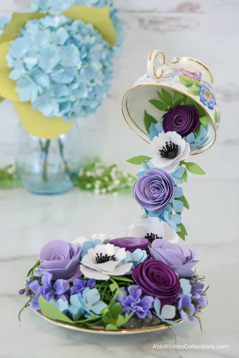 A white and gold tea cup with a floral design floats, tipped over in the air. A row of purple and white paper flowers spill out from the tea cup, cascading down into a saucer with paper flowers in shades of purple, blue, and white.