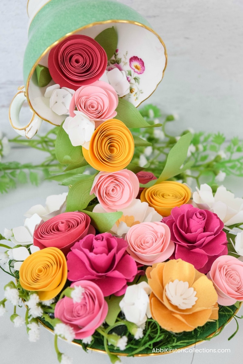 A floating tea cup pouring a stream of red, pink, and marigold paper rosette flowers onto a tea saucer. The colorful paper flowers on the saucer are filled in with bits of greenery and small white silk flowers.