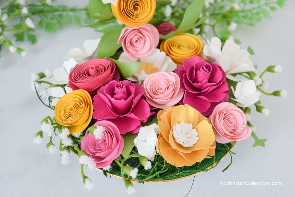 The saucer of the floating tea cup, filled with a colorful array of paper flowers, Light and dark pink, marigold, and white flowers fill the saucer, along with faux moss and greenery in the open spaces.