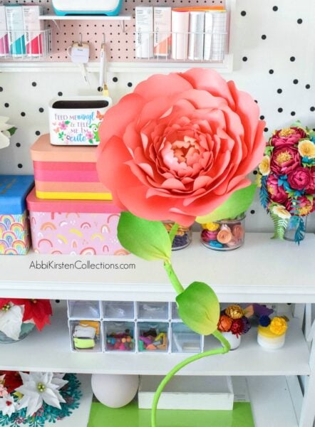 A red giant paper peony sits on a leafy, curvy free-standing stem. Behind the papercraft are shelves of organized craft supplies. 
