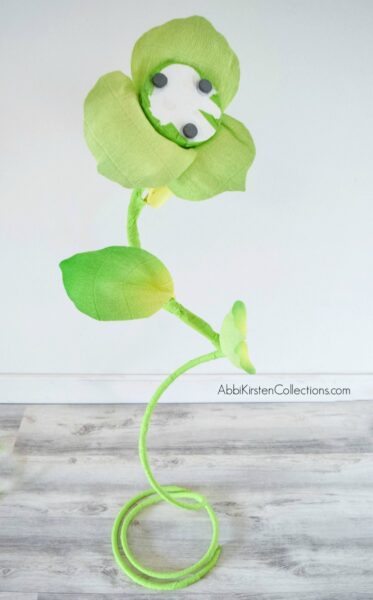 A free-standing green stem is in front of a white wall and a light brown wood floor. The copper wire is in the bottom and the flower will be placed on the magnets on top. 