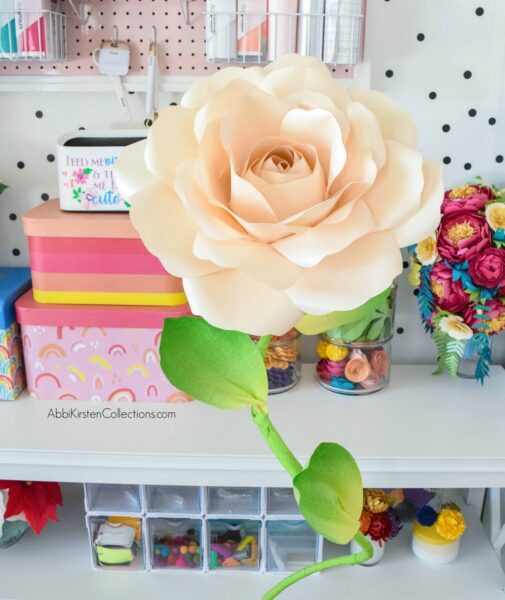 A large light pink paper rose with a giant green stem sits in front of organized craft shelves full of supplies. You can make these curved free-standing stems!