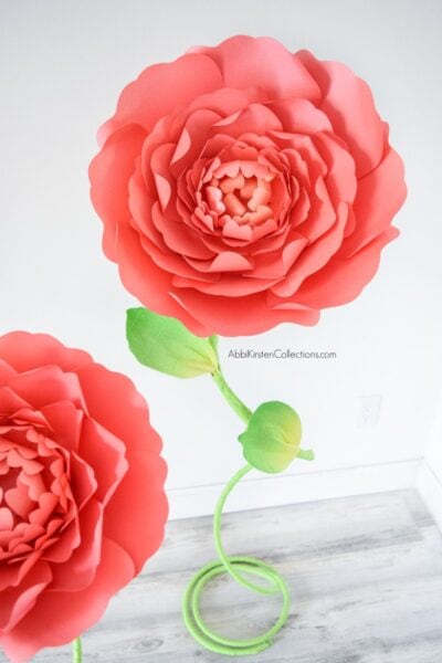 In the corner of a white room is a free-standing green stem and giant red paper flowers, with part of another red flower in the foreground. These DIY stems can be created using Abbi Kirsten's templates. 