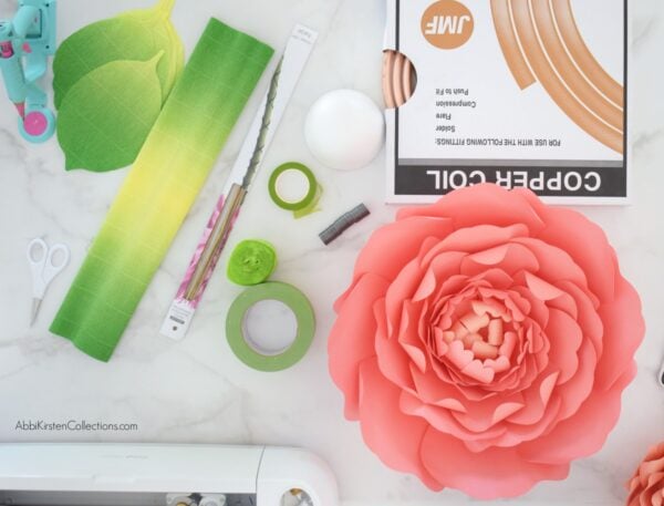 Pictured are the supplies needed to create free-standing green stems for large paper flowers. Copper coil, green leaves, a red giant flower, corsage tape and more lay on a marbled table. 