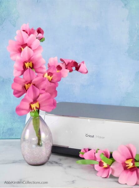 Pink DIY paper orchids fill a clear vase placed next to a Cricut machine in front of a swirled blue background. You don’t need a Cricut to create these flowers. 