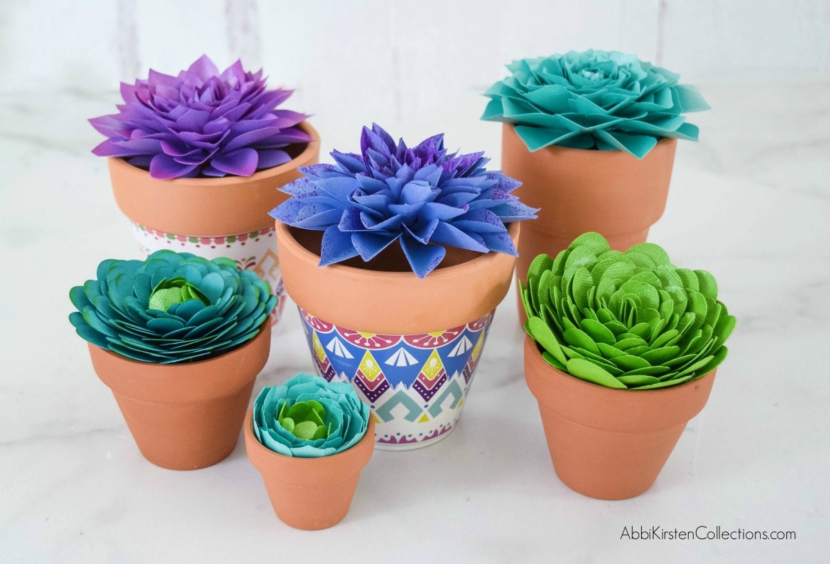 Six assorted terra cotta plant pots sit on a white floor. Each pot has a different colored paper succulent. You can learn how to make paper succulents with the tutorials provided by Abbi Kirsten. 