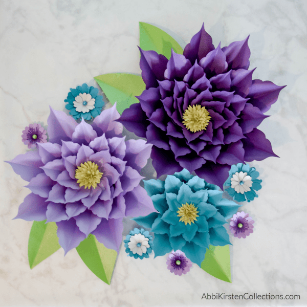 An overhead view of varying size paper amaryllis flowers in shades of purple and blue, surrounded by smaller blooms and green leaves.