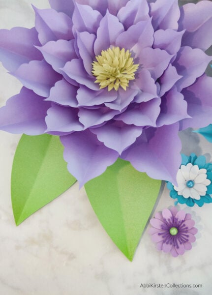 An overhead photo of a light purple paper amaryllis flower, with green leaves and smaller surrounding flowers.