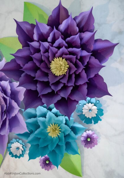A close-up of a giant purple and blue amaryllis paper flowers, surrounded by smaller flowers and green leaves.