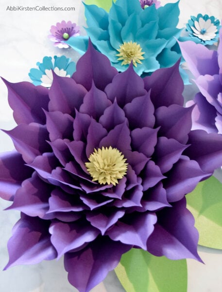 Giant, large, and small paper amaryllis flowers in shades of blue and purple with green leaves.