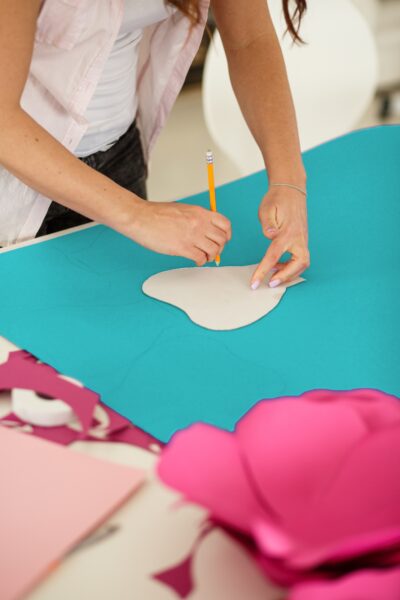 Abbi traces a heart-like shape on a large teal piece of cardstock. In the foreground is a giant dark pink flower. 