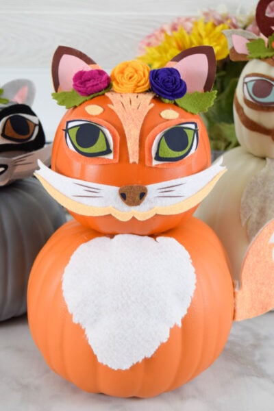 The orange fox is made from two pumpkins and wears a crown of three colorful flowers in dark pink, orange-yellow and deep blue. How to Make Woodland Animal Pumpkins for Easy and Fun Fall Decorating. Use felt and iron-on vinyl with your Cricut to craft a fox, deer and raccoon pumpkin decoration for your home!