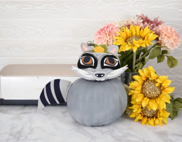 In front of a Cricut machine, sitting on a white and gray marble floor, is a gray raccoon made from felt, vinyl and two faux pumpkins. Next to the craft are bright yellow sunflowers. 