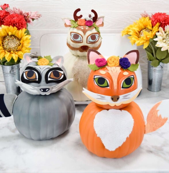 Amid colorful flowers in skinny metal vases sit three woodland pumpkin animals in gray, orange, and beige.  There is a raccoon, a fox, and a deer. These woodland animal pumpkins are fun and simple to make. 