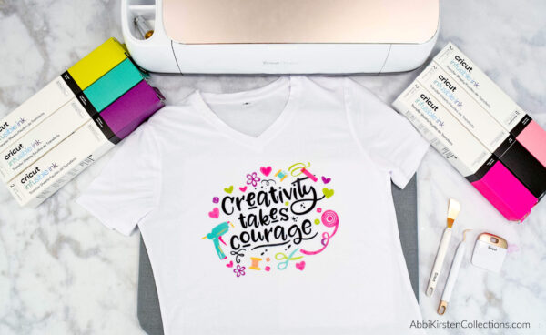 The Cricut Infusible Ink tutorial lists the supplies you need for successful projects. This picture shows infusible ink cartons, a printed t-shirt with the words "Creativity takes courage" printed on it, and some supplies needed to make your own Cricut crafts. 