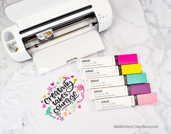 A photo of an open Cricut machine, cartons of infusible ink in multiple colors, and a white t-shirt with 
"Creativity take courage" written in black text surrounded by colorful craft supply illustrations. Cricut Infusible Ink directions suggest using light-colored shirts for the best, most vibrant results. 