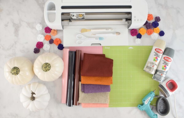 An overhead view of the supplies needed for this simple woodland animal pumpkin craft. In view are a Cricut machine, rolls of vinyl, faux pumpkins, felt and a glue gun. 