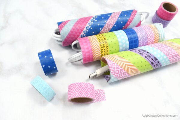 Upcycled cardboard tubes are covered in colorful washi tape stripes. The cardboard tubes are used as organizers for electrical cords and phone chargers.