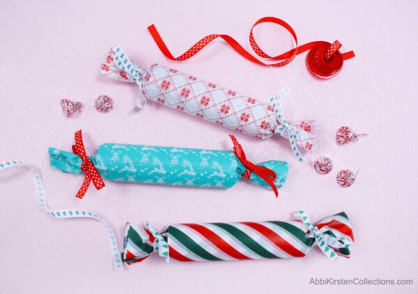 Recycled cardboard tubes make excellent holiday candy and small gift holders. These candy holders are shown on a pink background, with red and white ribbon. The tubes are wrapped in holiday tissue paper in red, green, and blue patterns.