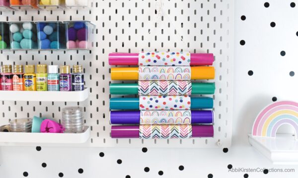 A craft space is organized with white shelves holding paint and white wallpaper with black polka dots. A DIY cardboard tube project is covered in bright rainbow scrapbook paper and hangs from the wall, holding an array of vibrant colored vinyl rolls. 
