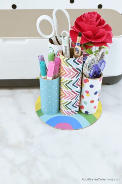 This pen organizer is made with upcycled cardboard tubes, each a different length, covered in washi tape and attached to a rainbow base. This desk organizer can hold pens, markers, scissors and more.