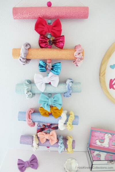A rainbow-colored hair bow organizer is made from upcycled cardboard tubes covered in faux glitter leather. The organizer holds scrunchies and bows in pastel colors and hangs on the white wall of a girl’s room. 