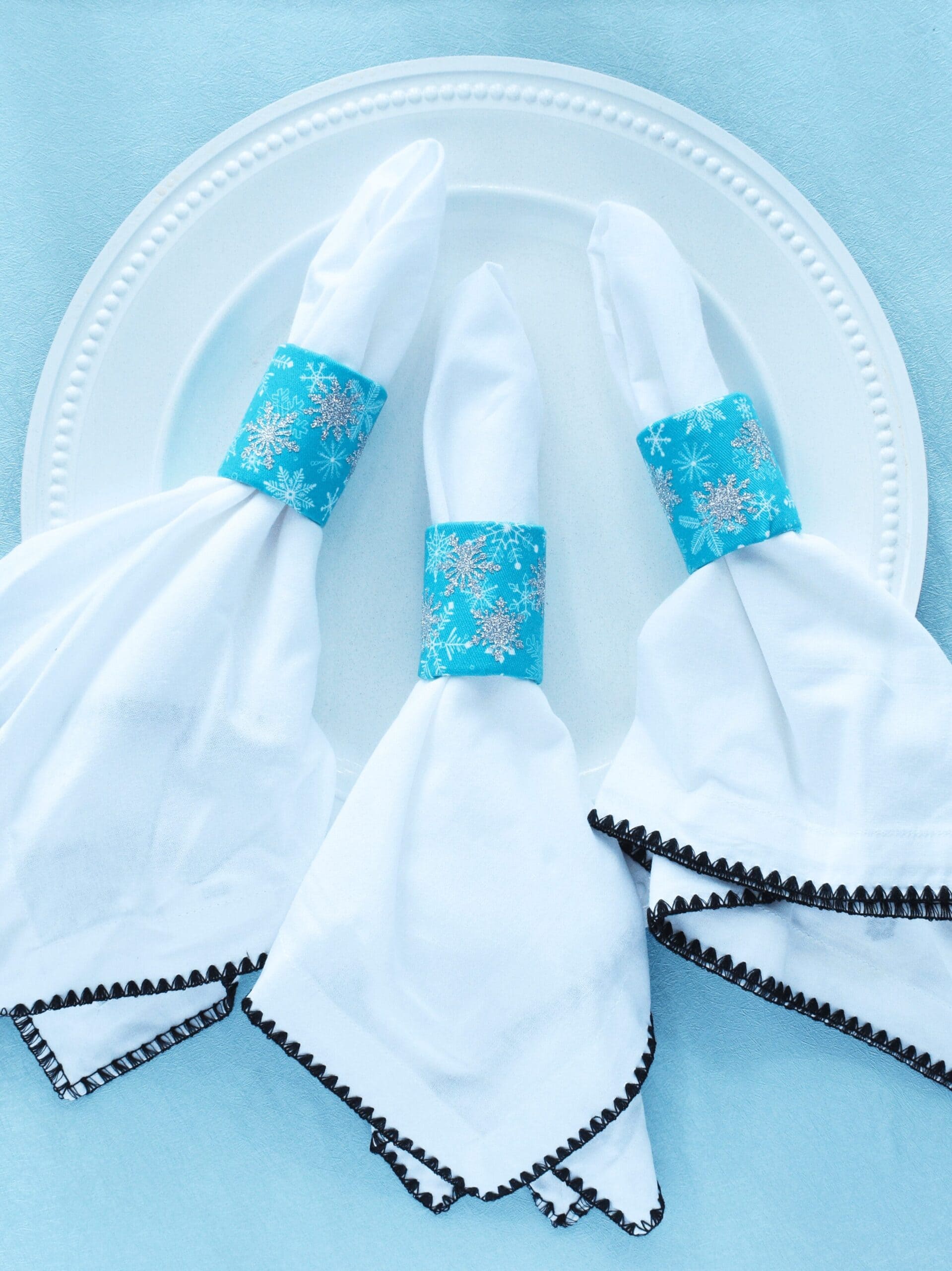 White napkins with black trim look festive and ready for a holiday tablescape thanks to upcycled cardboard tube napkin rings. The rings are covered in blue snowflake print fabric, and the napkins are against a white plate on a light blue background. 