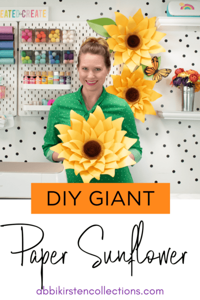 Abbi holds one of her giant yellow paper sunflowers in her craft room. The text says "Giant Paper Sunflower Tutorial by Abbi Kirsten."