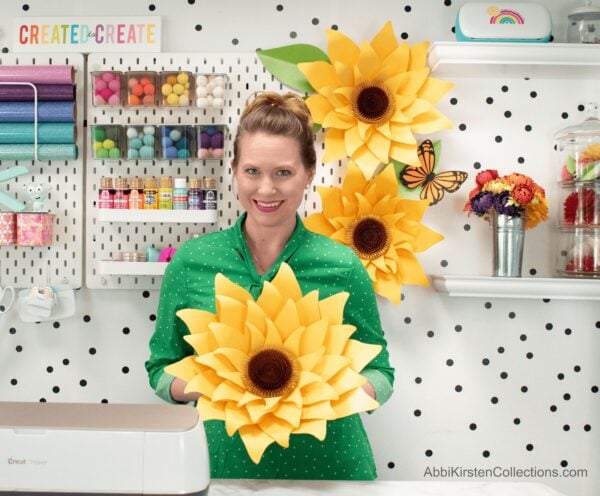 Abbi holds a giant yellow paper sunflower against her green shirt.  She is in her craft room with black and white polka dotted walls and shelves of craft supplies. 
