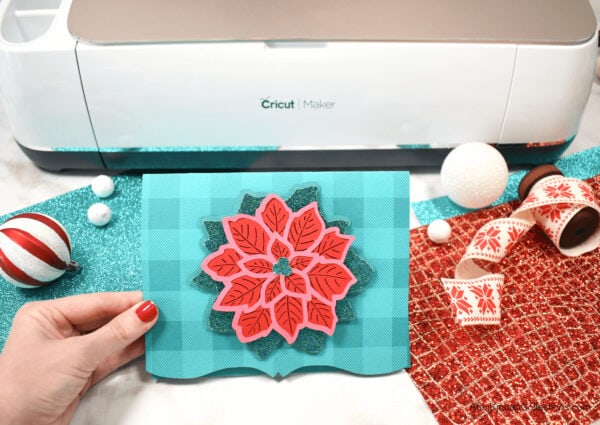 A gorgeous and festive Poinsettia Mandala Christmas Card held in front of a Cricut Maker cutting machine, surrounded by glitter craft paper and Christmas decorations.