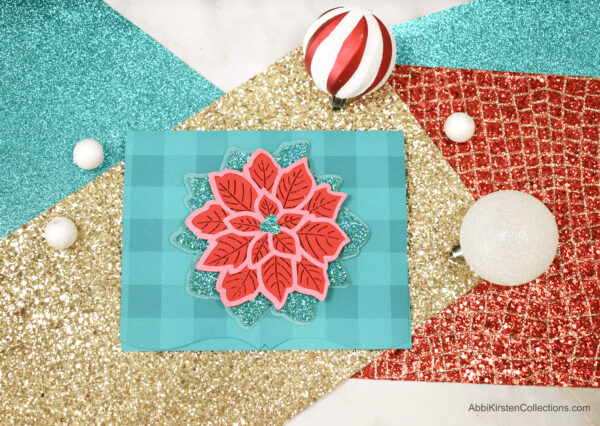 Green, red, and gold glitter card stock serves as a background for a fun and festive layered Poinsettia Mandala Christmas card, surrounded by Christmas ornaments.