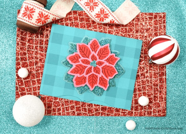 Image shows a festive layered Poinsettia Christmas mandala Christmas card is made with glitter craft paper, sitting on top of red and blue paper backgrounds, surrounded by Christmas ornaments and Christmas ribbon.