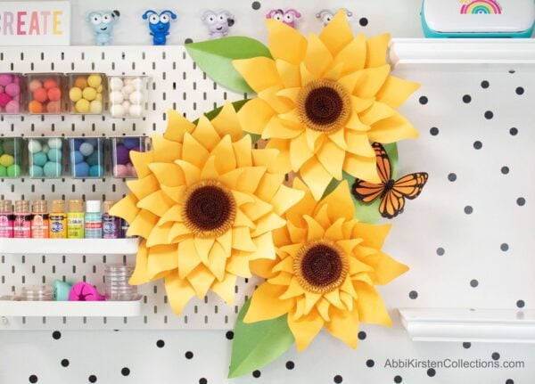 Giant yellow sunflowers with green leaves adorn a white and black polka dot wall with shelves holding colorful craft supplies like paint and pompoms. 
