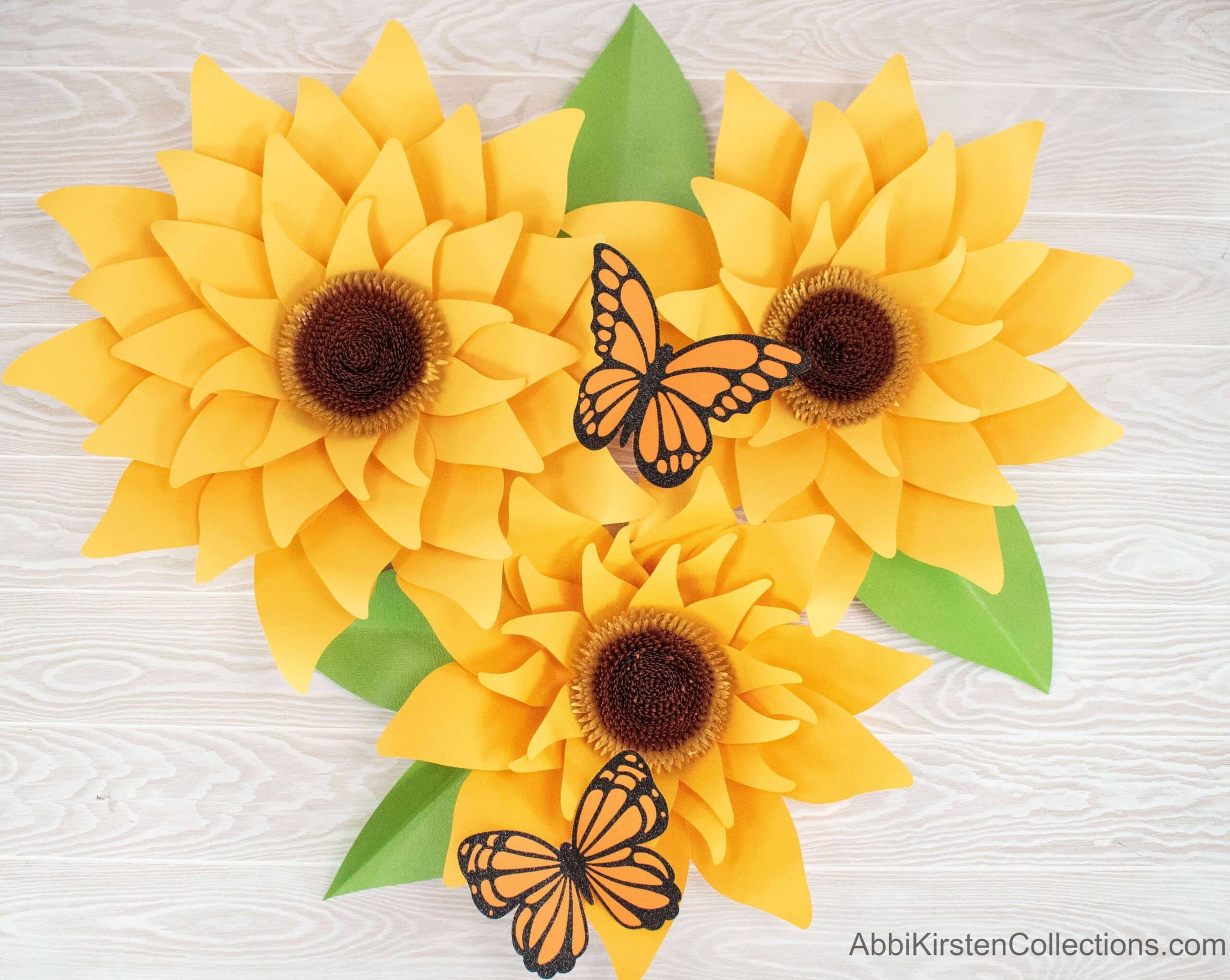 Three large paper yellow sunflowers viewed from above. Green leaves and Monarch butterflies adorn the flowers on a white and light gray wooden table. 
