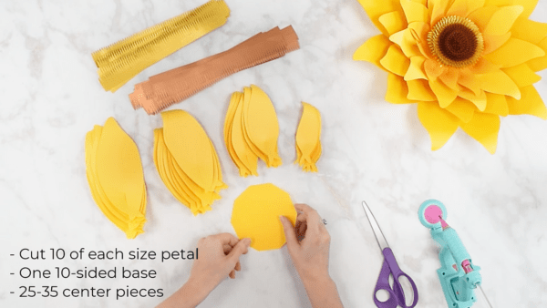 This overhead view of Abbi's white marbled craft surface shows the supplies needed to make a giant yellow sunflower. A completed sunflower is in the upper right corner and the petals and center cutouts are stacked on the left. 