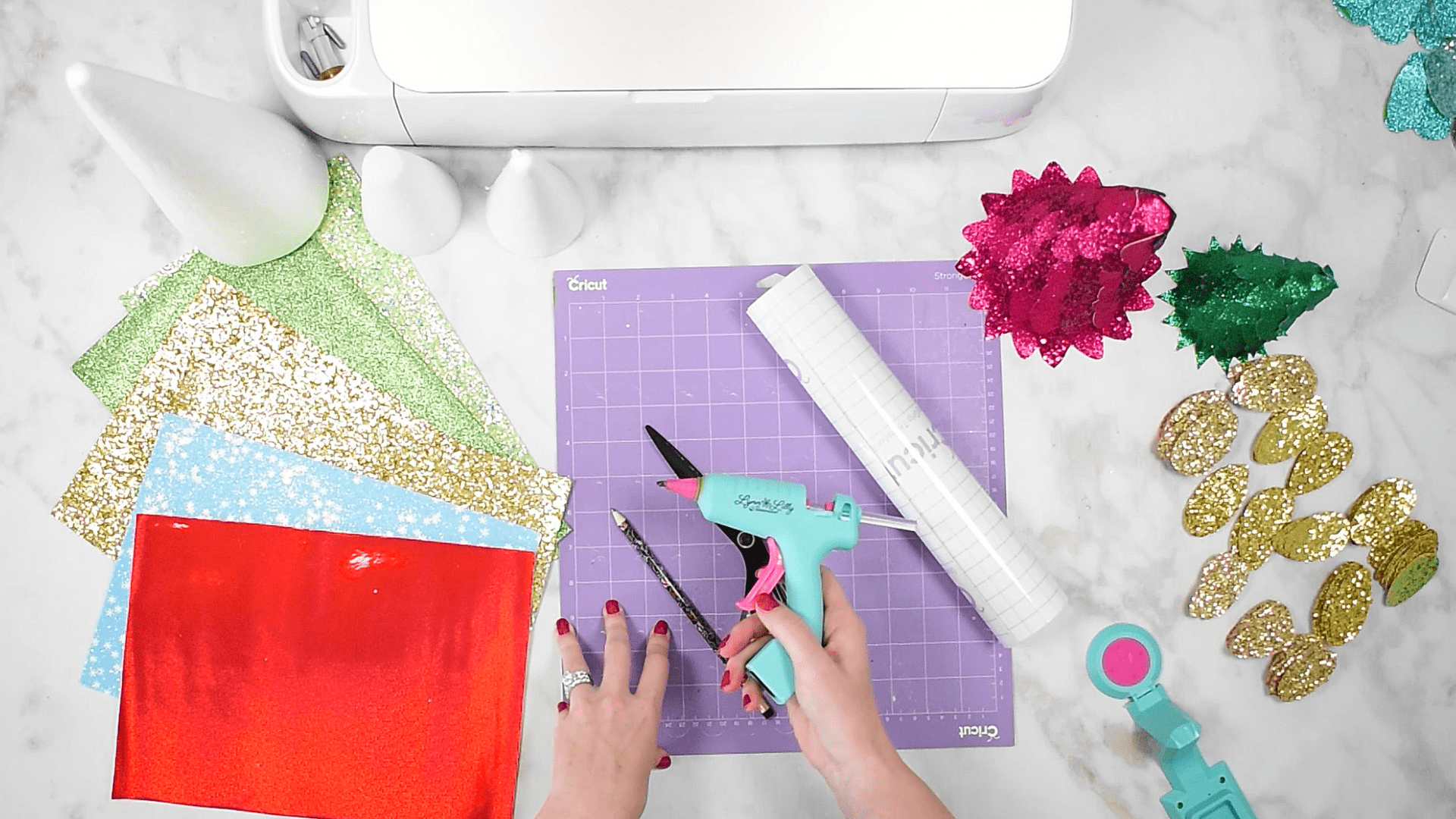 An overhead view of Abbi’s crafting station shows an array of craft supplies to make faux leather Christmas trees – styrofoam cones, sheets of glittery faux leather, and a Cricut cutting machine and accessories. Abbi’s hands are pictured holding a blue and pink glue gun.