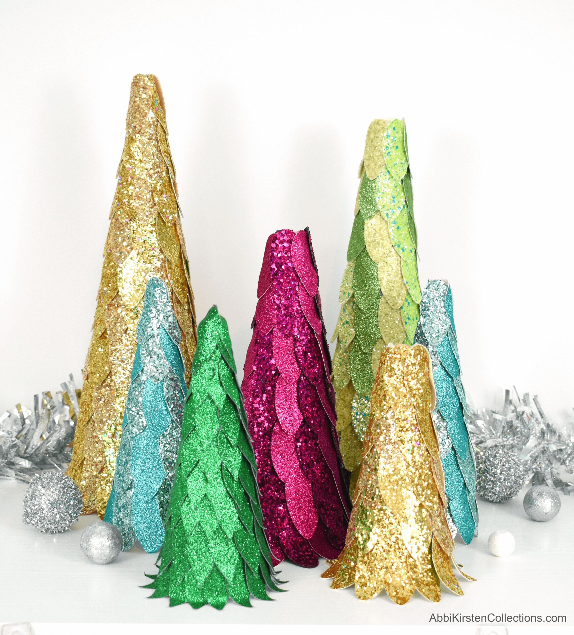 Seven DIY faux leather Christmas tree crafts stand together in front of decorative silver garland and Christmas ornaments. Each tree is a different height and created with layers of sparkling faux leather petals in festive gold, green, blue, silver, green, and pink. 