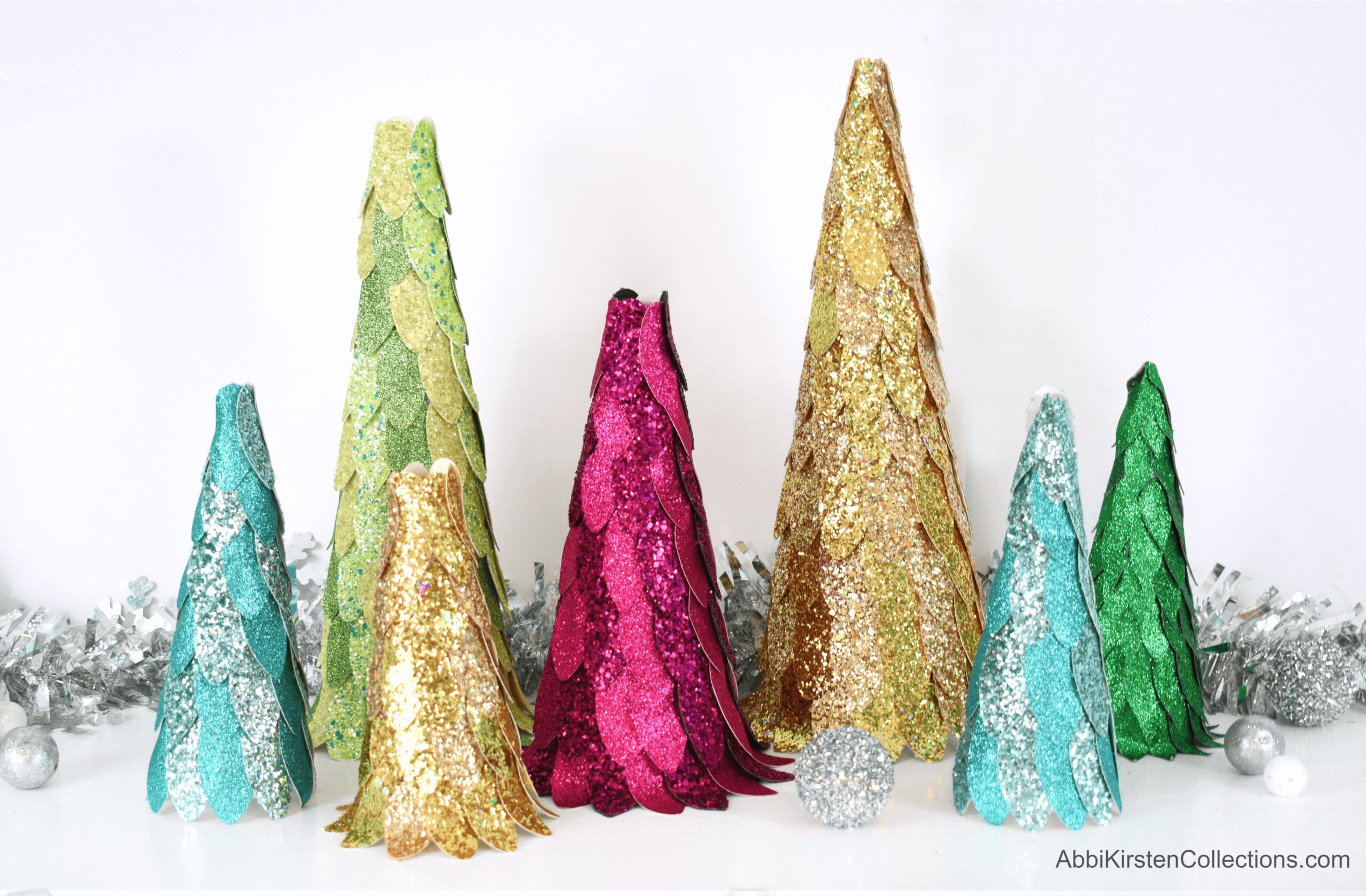 Seven sparkling faux glitter leather Christmas trees of varying heights sit on a white table in front of a silver garland. The trees are made from blue, silver, green, gold, and pink faux leather.