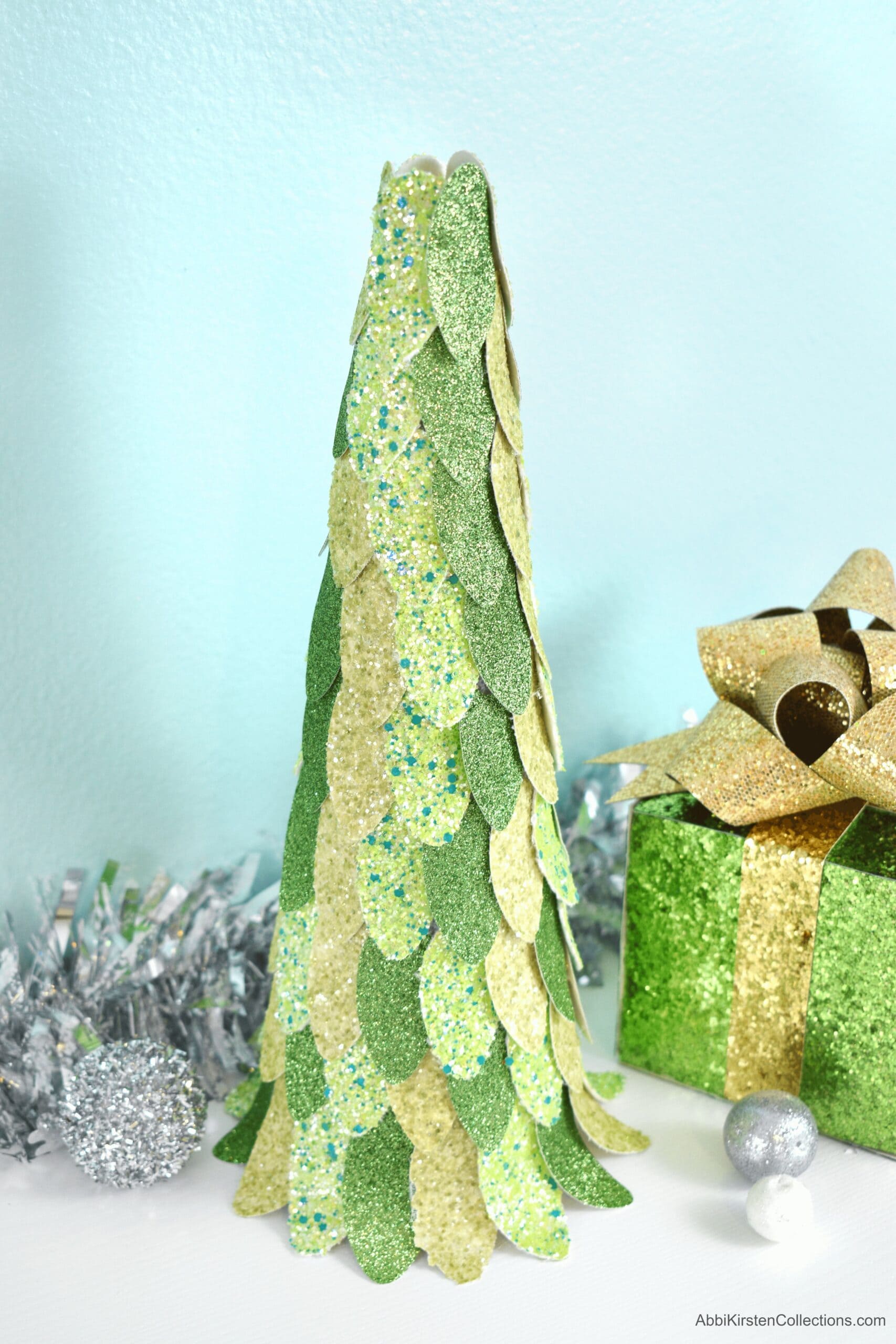 A tall, glittery green Christmas tree is made from layers of faux leather petals attached to a simple styrofoam crafting cone. In the background are a light blue backdrop, silver garland, and a green and gold wrapped present.