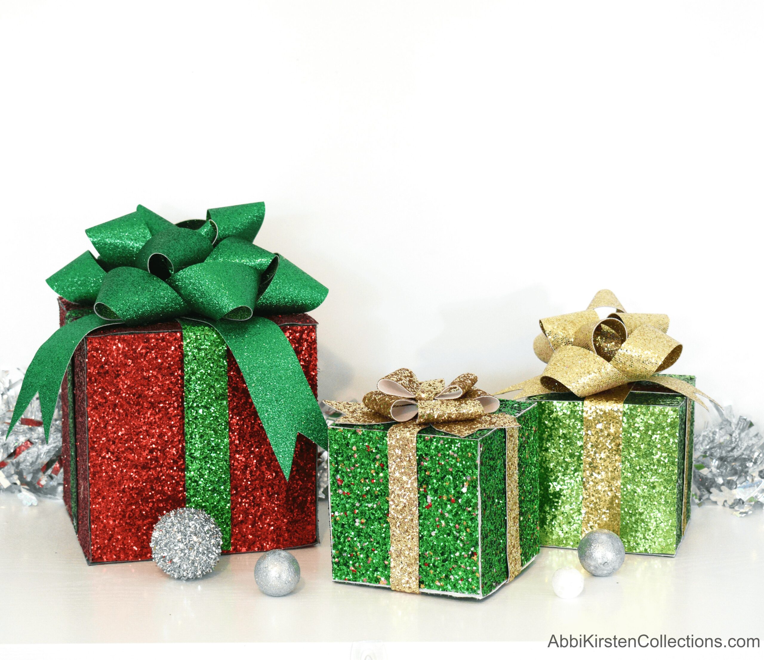 Three decorative gift boxes of different sizes, covered in red and green faux glittery leather sheets and topped with green and silver bows. Silver ornaments and tinsel surround the presents on the floor.