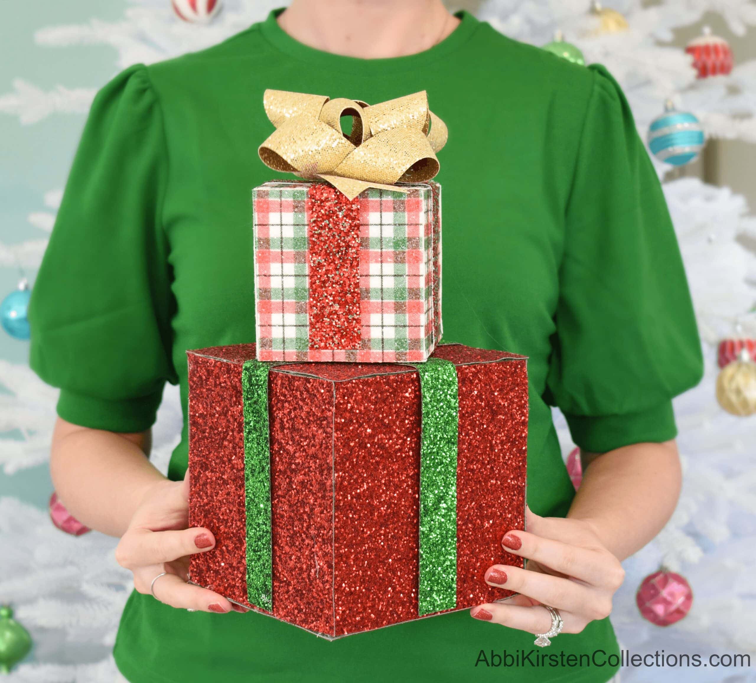 A woman wearing a short-sleeved green shirt holds two stacked decorative gift boxes. The larger box is red with green ribbon, and the top box is a red, white, and green plaid with a gold ribbon.
