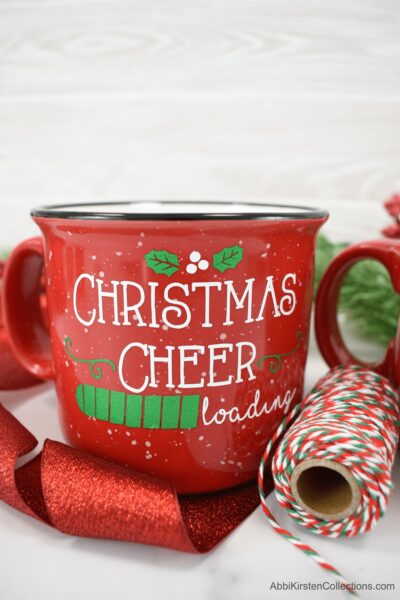 A red Christmas mug with the words "Christmas cheer loading" applied in permanent vinyl with Cricut. A red glitter ribbon and red and green twine surround this last minute Christmas gift. 